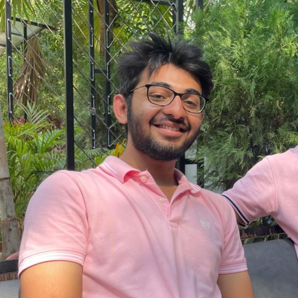 A smiling young man with glasses and a beard takes a selfie portrait outdoors, surrounded by lush green foliage and wearing a pink polo shirt.