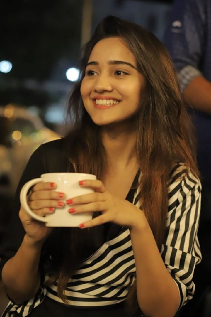  A smiling young woman holding a mug, wearing a striped top, with string lights in the background, likely at a cafe or restaurant.