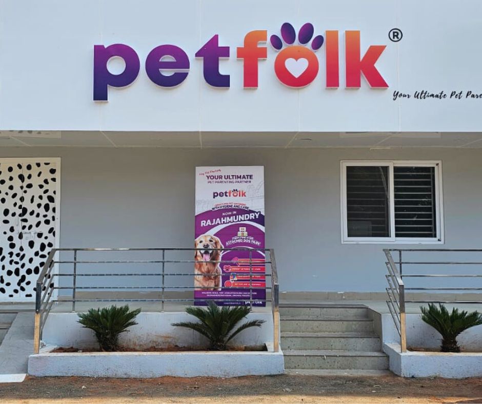 Petfolk storefront with a bright sign and logo above the entrance. The logo features 'petfolk' in bold, colorful letters with a paw print design incorporated into the letter 'o.' Below the sign, there is a banner advertising pet services in Rajahmundry, featuring a picture of a happy Golden Retriever. The entrance has a ramp and stairs leading up to it, with small green plants on either side.