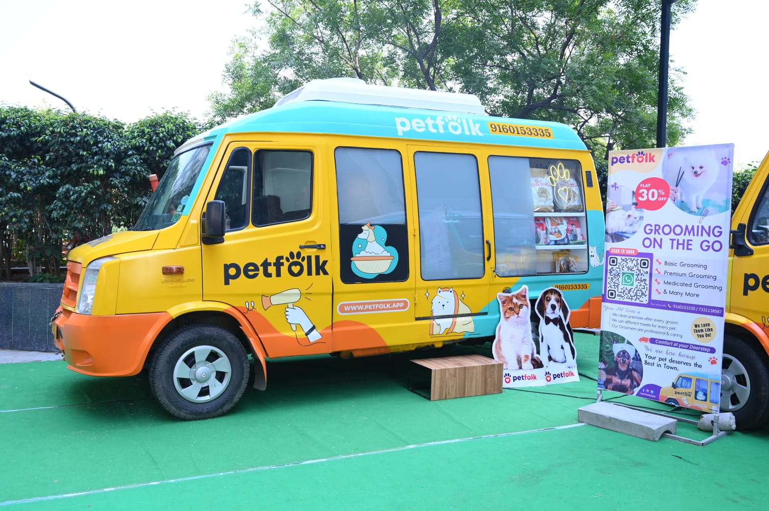 Petfolk mobile grooming van parked on a green mat, offering 'Grooming on the Go' services. The yellow and blue van is branded with Petfolk's logo and features illustrations of pets. A nearby sign promotes a 30% discount on grooming services, listing options like basic, premium, and medicated grooming. Cutouts of cats and dogs are displayed next to the sign, attracting attention to the mobile grooming service.