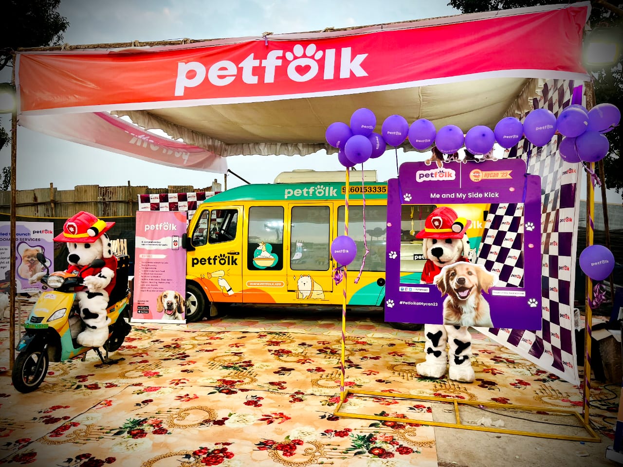 Petfolk promotional event setup with a large tent displaying the Petfolk logo. Under the tent, a costumed character dressed as a Dalmatian firefighter rides a scooter, while another poses with a photo frame alongside a Golden Retriever image. A brightly colored Petfolk van, adorned with pet care graphics, is parked behind them. Purple balloons with Petfolk branding and various banners promoting pet services are displayed around the area.